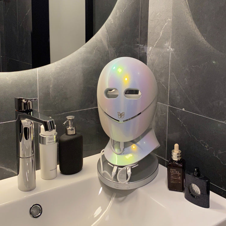 Still image of CHOUCHOU TOKYO LED mask displayed in a bathroom among perfume bottles and skincare products, creating an elegant and realistic daily routine setting.